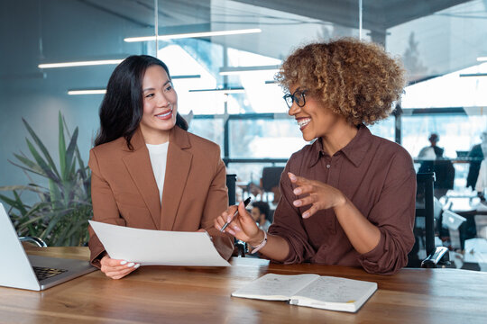 Two businesswomen collaborating at modern office desk smiling during meeting with laptop and documents. Multiracial women talking working together. Teamwork and professional communication at workplace