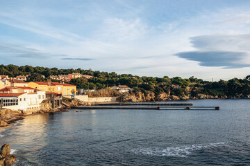 Naklejka premium Hyeres, France - December 7, 2025: Coastal view of Port de la Tour Fondue harbor with Mediterranean houses, rocky shoreline and calm sea under soft winter light