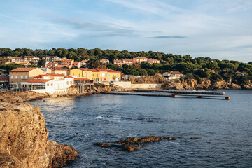 Fototapeta premium Hyeres, France - December 7, 2025: Coastal view of Port de la Tour Fondue harbor with Mediterranean houses, rocky shoreline and calm sea under soft winter light