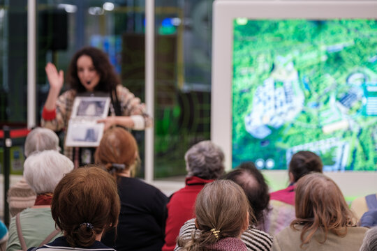Group of older adults listening to presenter during guided tour, digital map screen in background, community education, social engagement, learning.