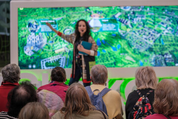 Fototapeta premium Female speaker presents urban planning map to seated audience at exhibition, seniors listening closely, educational tour, technology display, community engagement.