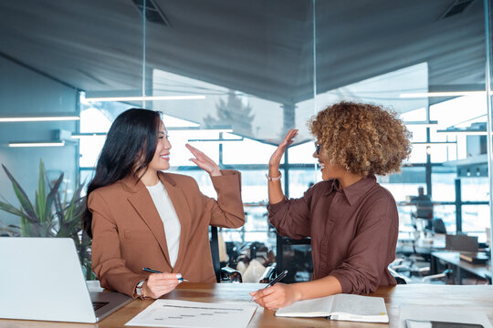 Two diverse businesswomen in modern office giving high five over desk, smiling during teamwork meeting; laptop and documents on table, open-plan workspace. Team of women collaborating