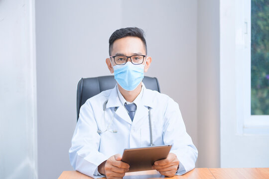 Portrait of a male doctor wearing a face mask and laboratory coat sitting at a desk