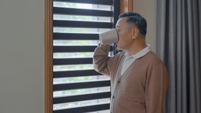 Lonely elderly man standing drink hot tea or coffee and looking at the window closed with curtain for thinking or nostalgia. Retirement, dementia, oldness, disability, nursing home concept