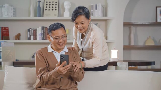 Smiling elderly couple wife husband make video call conference using smart phone sit on couch at home. Old man woman holding smartphone have funny conversation with friends, relatives or grandchildren