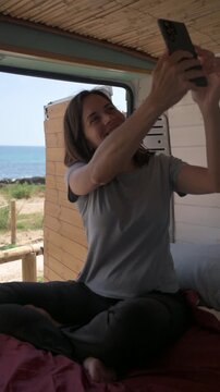 Young woman sitting on a bed inside her camper van taking a selfie with the sea in the background
