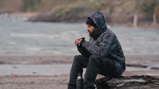 A man in a hooded jacket drinks mate while seated on a log by a windswept lakeshore. The scene captures a contemplative moment in a rugged outdoor setting. Suitable for travel content or lifestyle blo