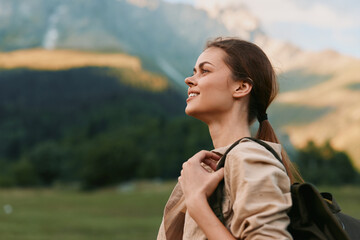 Obraz premium Woman traveler smiles while wearing a backpack in a meadow, with distant mountains, embodying outdoor lifestyle and exploration on a bright calm day. Scenic backdrop boosts mood.