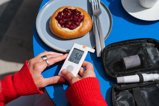Overhead personal perspective of a woman sitting at a table at an outdoor cafe using a glucometer to measure her blood sugar before eating a cherry brioche bun