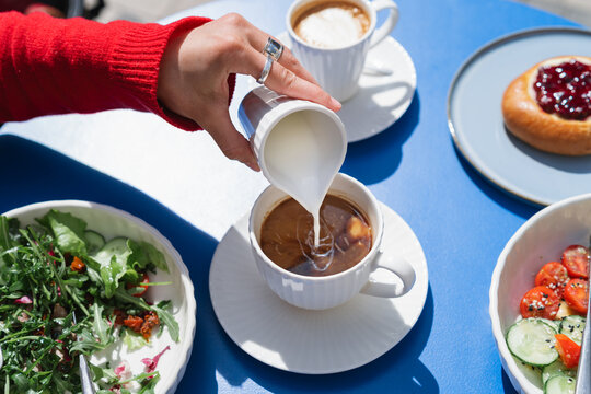 Close-up overhead view of a woman's hand pouring milk into a cup of black coffee before eating a beef salad, quinoa salad and cherry brioche bun