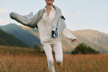 Naklejka premium Young woman in white knit sweater and pants joyfully runs through a grassy field with mountains in the background on a clear day.