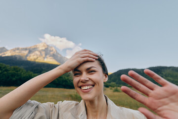 Naklejka premium Cheerful woman outdoors, smiling with hand on head, sunlit moment, relaxed posture, natural colors, meadow and mountains in the background, gentle breeze, blue sky