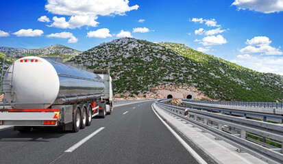 Fototapeta premium A large tanker truck travels along a modern highway winding through a mountainous landscape toward a tunnel under a bright blue sky.