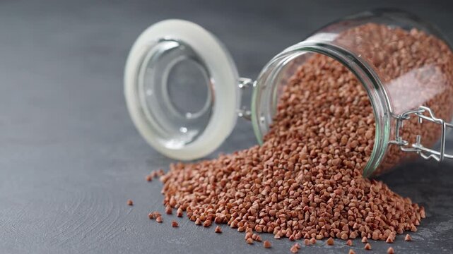 Organic buckwheat spill out of a glass storage jar on a dark stone table. buckwheat spill out of a glass storage jar a dark table. Raw buckwheat are scattered out of glass jar.