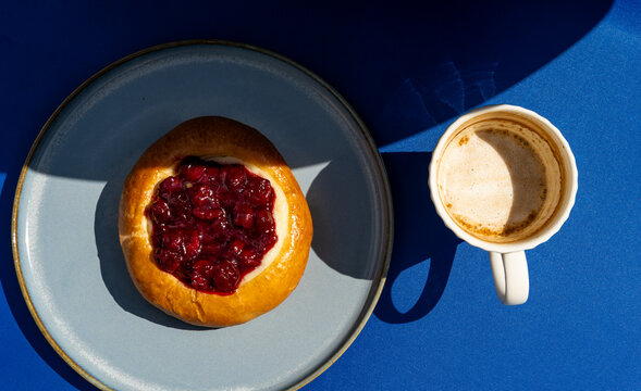 Overhead view of a cherry brioche bun and a cappuccino on a blue background on a sunny day