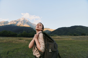 Naklejka premium Young woman with a backpack gazes upward in an open meadow, mountains in the distance. Outdoor lifestyle, exploration, and self discovery through hiking journey.