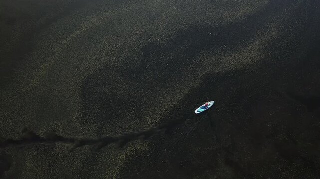 Minimal aerial view of a woman paddleboarding on dark water with subtle golden light reflections. Top-down drone perspective with strong negative space and abstract texture of water surface