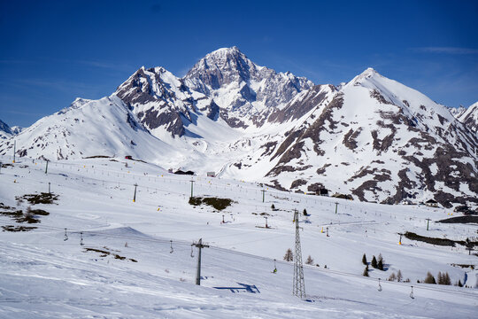 Aerial view of the snow-covered peaks of the Mont Blanc massif and ski slopes with lift infrastructure under a clear blue sky in La Thuile, Valle d'Aosta, Italy.