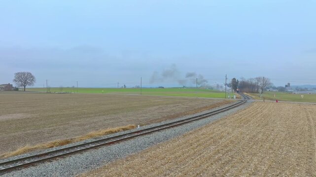 A train moves along a railway track through open fields. Smoke rises in the background. The sky is grey with clouds above. The rural area shows a flat landscape.