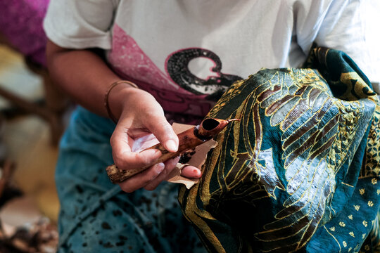 Close-up of an artisan using a canting tool to apply hot wax onto batik fabric. Capturing the intricate traditional hand-drawn process with brown wax on dark patterned textile.