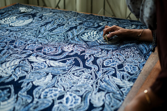 Perspective view of an artisan's hand guiding a canting over white fabric to create wax-resist motifs. Highlighting the cultural heritage of Javanese textile art and production.