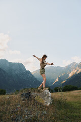 Naklejka premium Young woman balancing on a large rock in a mountain meadow during daytime. Outdoor lifestyle concept with nature, adventure, and freedom in a scenic landscape.
