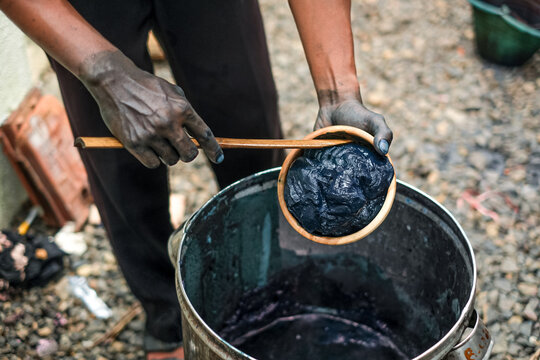 Mid-section view of a craftsman with hands stained dark blue holding a bowl of thick indigo paste over a deep metal processing tank.