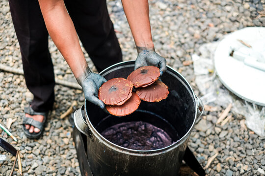 Perspective view of a craftsman with hands stained blue holding molded palm sugar discs right above a deep metal tank filled with natural indigo dye mixture.