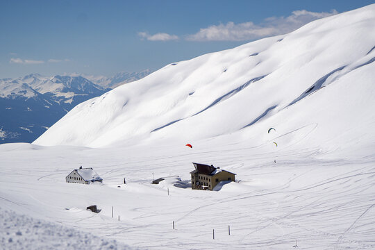 Aerial view of Colle del Piccolo San Bernardo with snow-covered mountain huts and paragliders under a clear blue sky in La Thuile, Valle d'Aosta, Italy.