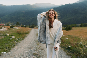 Fototapeta premium Smiling woman in cozy sweater and shawl walking on rural path with mountains in background. Outdoor lifestyle, nature, happiness, and casual fashion concept.