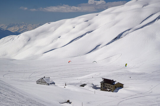 Aerial view of Colle del Piccolo San Bernardo with snow-covered slopes, mountain huts, and snowkiters gliding over the white terrain under a clear blue sky in La Thuile, Valle d'Aosta, Italy.