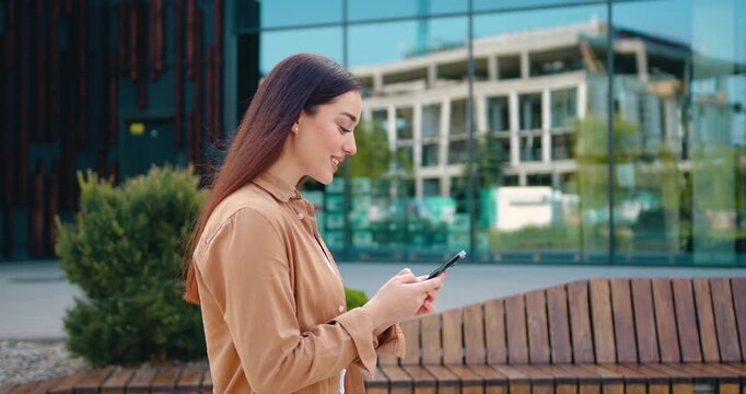 Side view of a smiling young woman with long hair in casual wear walking along a city street near a modern office building, using her smartphone to reply to messages.