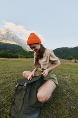 Naklejka premium A woman outdoors in a bright field with an orange beanie bows over a backpack, preparing gear in a scenic mountain landscape for an adventurous lifestyle moment.