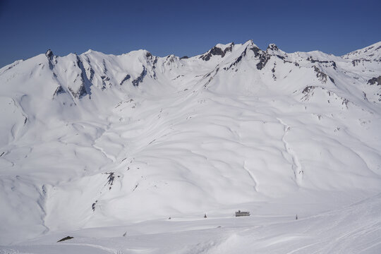 Aerial view of Colle del Piccolo San Bernardo with snow-covered mountain peaks and alpine slopes under a clear blue sky in La Thuile, Valle d'Aosta, Italy.