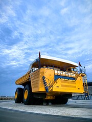 Low angle view of a giant yellow mining dump truck with massive tires, blue metal stairs and safety railings against dramatic cloudy blue sky. Heavy industrial mining vehicle concept. © Алина Неборская