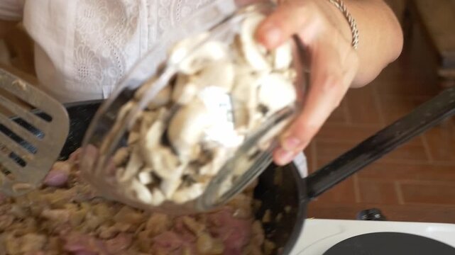 Medium shot of a woman adding sliced ​​mushrooms and red wine to a pan with meat. Natural interior lighting. Concept of gourmet home cooking, artisanal recipe, and culinary preparation.