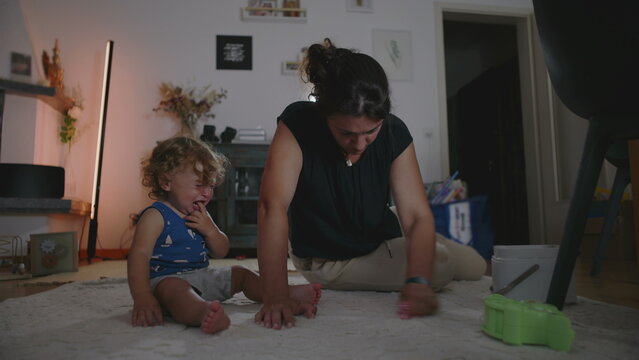 Mother rubbing and cleaning carpet to remove a stain while toddler cries beside her seeking attention, candid indoor family moment showing daily parenting challenges