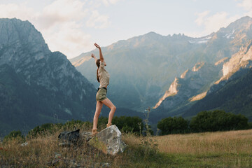 Naklejka premium Young woman in casual clothes stands on a rock with arms raised in a mountain field, enjoying freedom and nature during a sunny day.