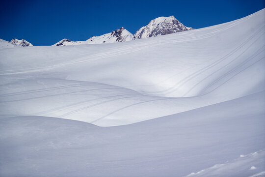 Aerial view of the snow-covered slopes of Colle del Piccolo San Bernardo with ski tracks and mountain peaks under a clear blue sky in La Thuile, Valle d'Aosta, Italy.