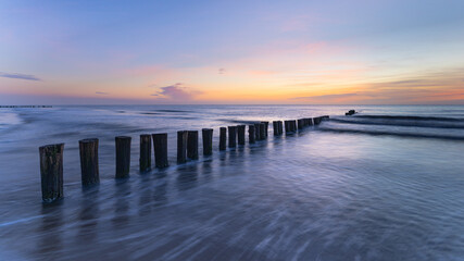 Fototapeta premium Wooden groynes in the sea at sunset – North Sea coast, Schoorl, the Netherlands. A line of weathered wooden groynes stretches from the foreground into the sea.