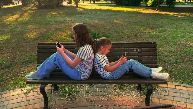 Sisters sitting separately on a bench, Modern childhood in the park, Digital engagement among kids, Girls disconnected while being together, Sibling bonding with screens, Summer day with technology