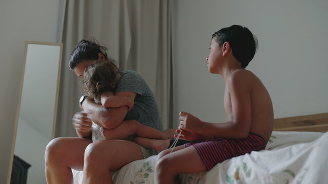 Mother comforting crying toddler in emotional embrace while older brother sits quietly on bed, family stress, parenting challenges, overwhelmed mother, mental and emotional fatigue