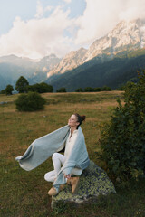 Naklejka premium Young woman wrapped in a blanket sitting on a rock in a mountain meadow. Relaxed lifestyle concept with scenic nature background and soft natural light.