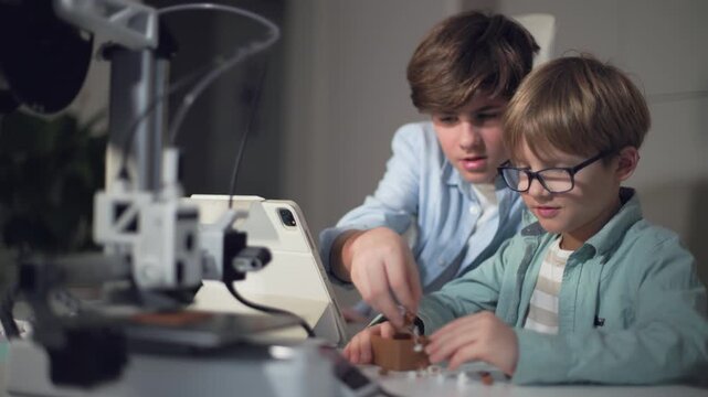 Two boys at a table with a working 3D printer, observing and operating it