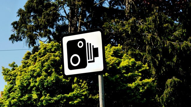 Speed camera sign, UK traffic enforcement symbol, police speed trap notification on white road sign with green trees