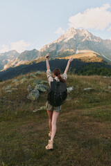 Naklejka premium Smiling woman with a backpack raising her arms in a grassy meadow, mountains in the background. Outdoor lifestyle, adventure, and personal achievement. Bright natural light.