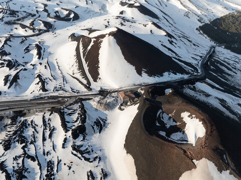 Aerial view of the snow-covered volcanic craters on the south side of Mount Etna with winding roads and dark volcanic ash in Nicolosi, Sicilia, Italy.