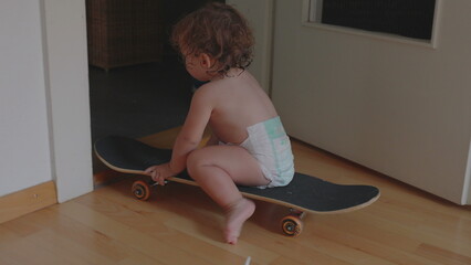 Toddler sitting on skateboard wearing diaper indoors, exploring balance and movement, playful childhood moment of curiosity and independence at home © Marco