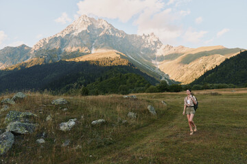 Naklejka premium Hiking in a meadow with mountains in the distance, wearing a backpack and trekking gear. A scenic landscape captures outdoor lifestyle, nature, and calm adventure.