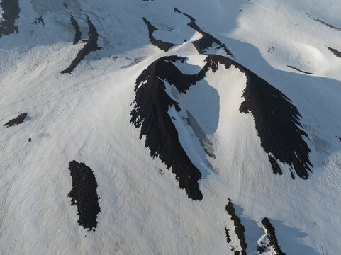 Aerial view of the snow-covered volcanic craters on the south side of Mount Etna with dark volcanic rock contrasting against the white snow in Nicolosi, Sicilia, Italy.